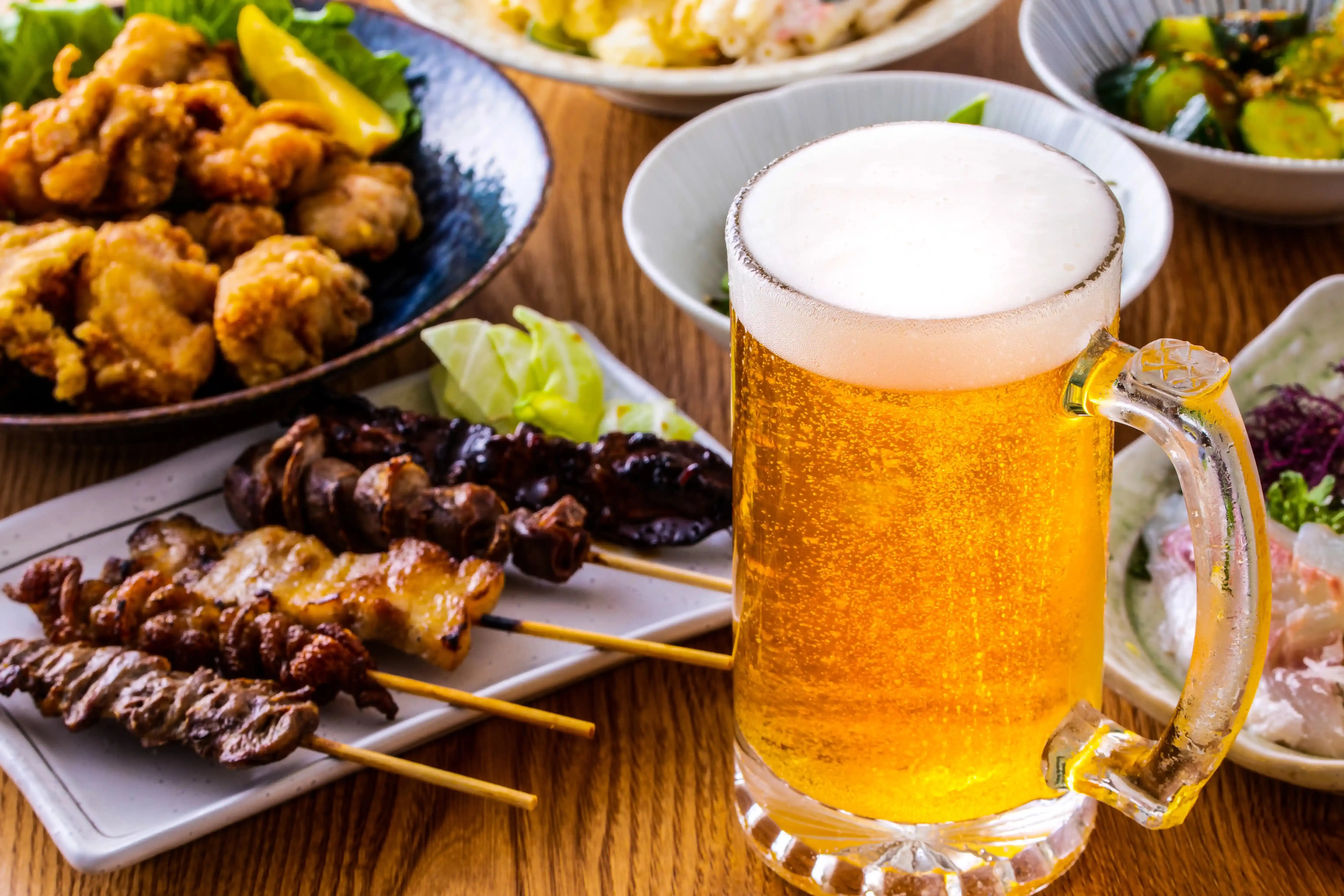 Caneca de cerveja clara e espumosa sobre uma mesa de madeira, rodeada por espetinhos de carne grelhada e frango frito, ilustrando a dúvida se a cerveja sem álcool faz mal para a saúde.