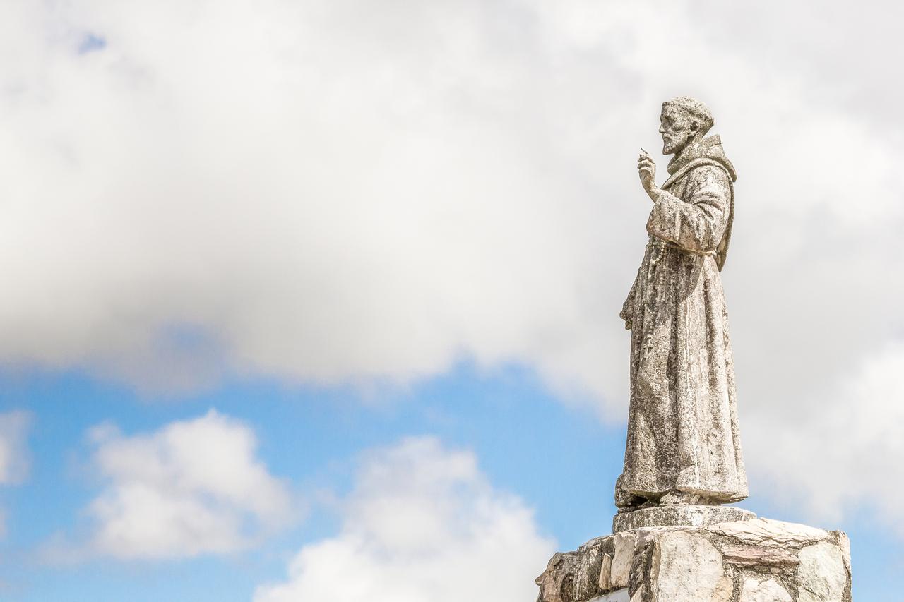 Estátua de São Francisco de Assis em pedra, com céu nublado ao fundo, simbolizando paz e espiritualidade.