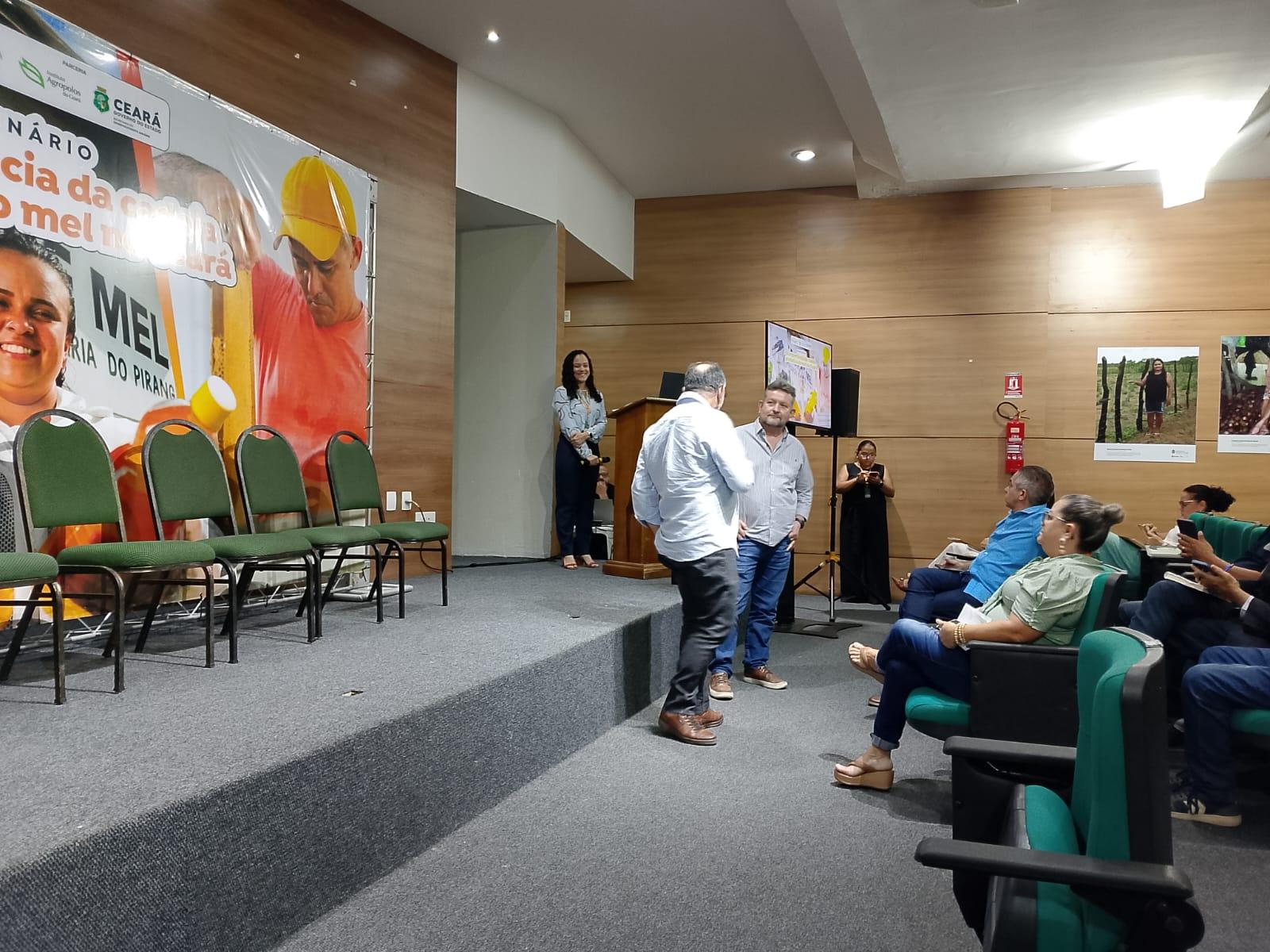 Imagem mostra auditório com pessoas próximas a um palco. Há um banner na parede desse auditório com a logo do Governo do Ceará.