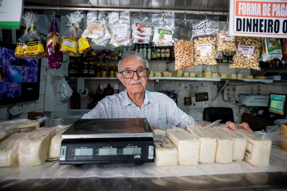 raimundo do queijo, um idoso, de cabelo branco, óculos e bigode, atrás do balção de sua mercearia, com uma balança e queijos ao lado