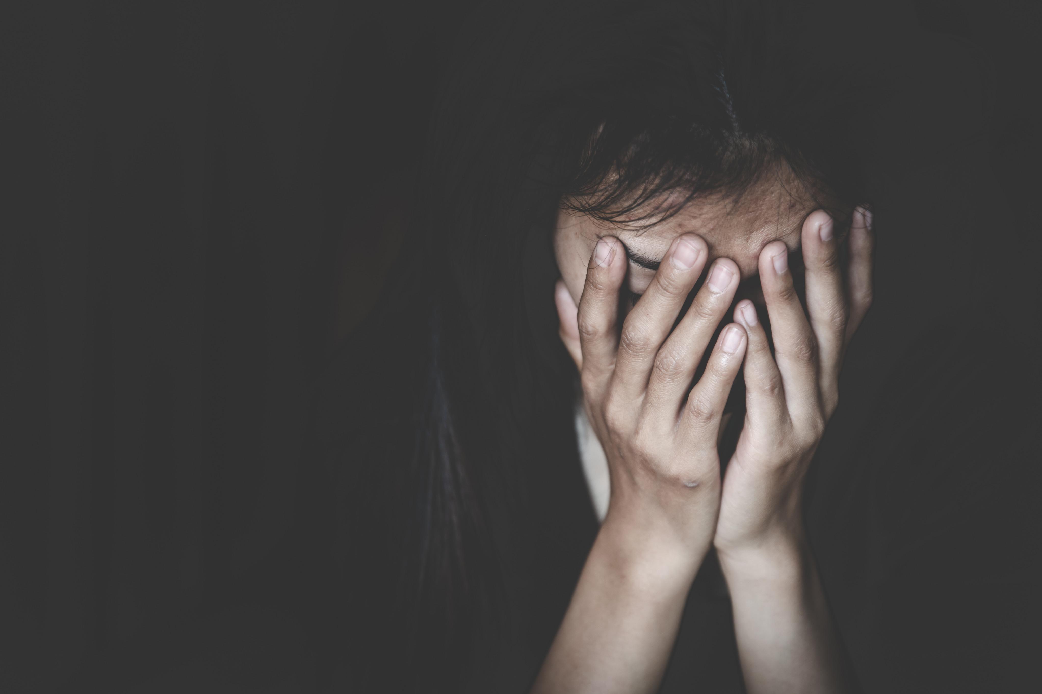 Uma fotografia em baixo-contraste e com iluminação dramática. A imagem mostra uma pessoa, possivelmente uma adolescente ou jovem, sentada no escuro. Ela está com as mãos cobrindo o rosto, na altura dos olhos e nariz, em uma expressão de angústia, tristeza, choro ou desespero. A iluminação foca apenas nas mãos e parte do rosto e cabelo, enquanto o restante do fundo é completamente escuro. A foto evoca sentimentos de dor, depressão ou isolamento.