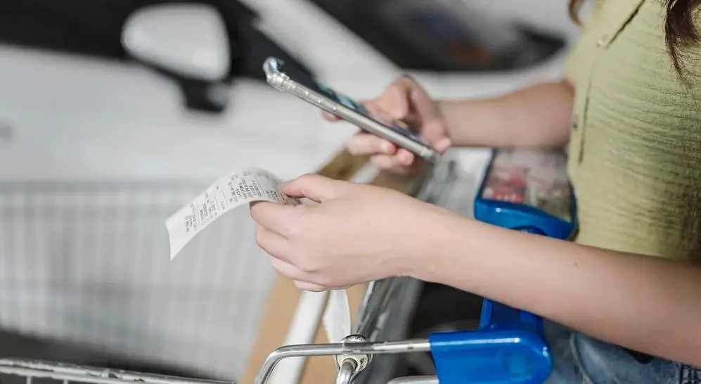 Mulher segura nota fiscal e celular em supermercado. Imagem usada em matéria sobre intoxicação por metanol.