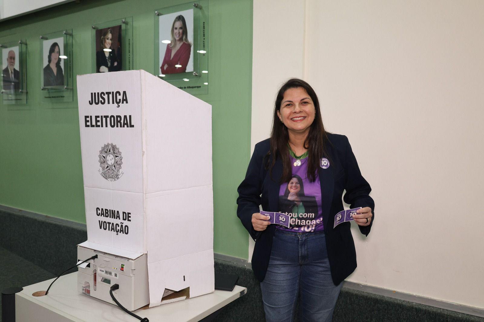 Elizabeth Chagas sorri ao lado de uma cabine de votação da Justiça Eleitoral. Ela veste camiseta roxa com sua própria imagem estampada e a frase 