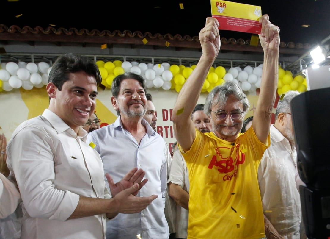 Grupo de político em um palco durante evento do PSB. No centro, Acilon vestindo camiseta amarela com a inscrição “PSB 40 Ceará” aplaude sorrindo, cercado por apoiadores que também batem palmas e tiram fotos com celulares. Ao fundo, há uma decoração com balões brancos e amarelos e um painel com símbolos gráficos e o mapa do Ceará. A atmosfera é festiva e de celebração.