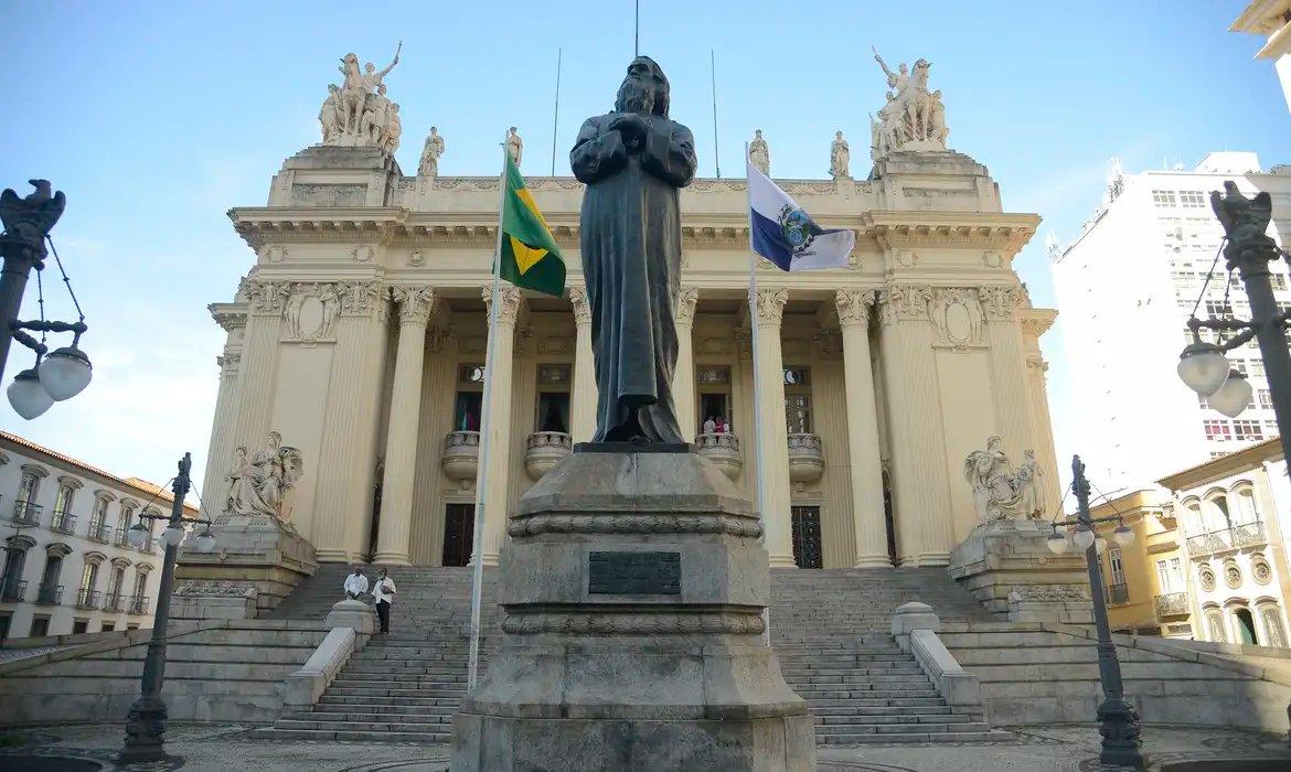 Imagem que representa a ALERJ (Assembleia Legislativa do Rio de Janeiro). Prédio de concreto bege com arquitetura atrás da estátua de  Tiradentes, uma escultura em bronze.