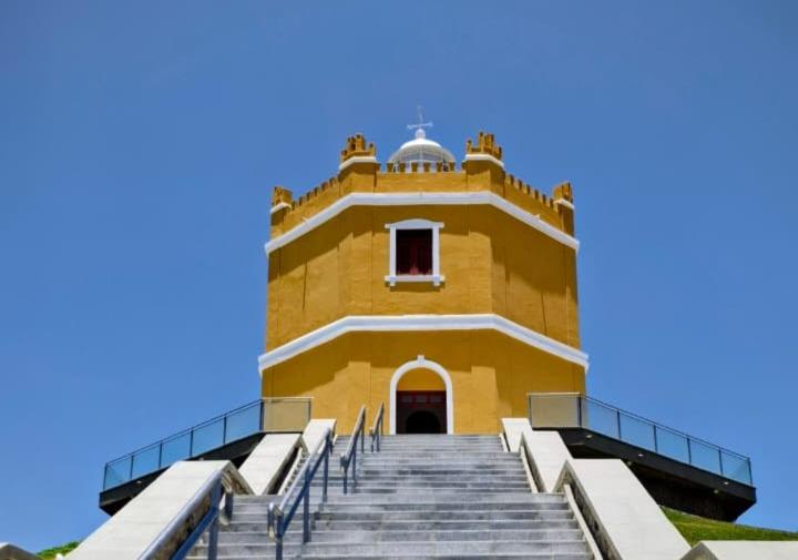 Escadaria central em pedra leva ao Farol Velho do Mucuripe, prédio histórico octogonal de cor amarela com detalhes brancos, janelas vermelhas e cúpula branca no topo, sob céu azul claro.