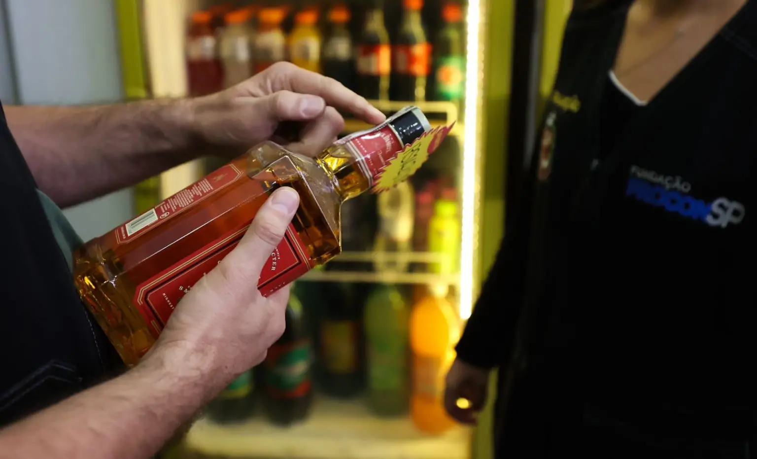 Pessoa segura uma garrafa de bebida alcoólica com rótulo vermelho diante de um refrigerador com garrafas de refrigerante; ao lado, agente da Fundação Procon-SP acompanha a fiscalização.