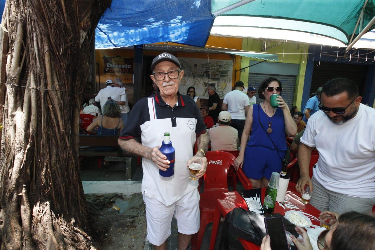 Foto de Raimundo do Queijo segurando cerveja. Ao redor dele estão pessoas bebendo cerveja e sentadas em uma mesa. Duas pessoas estão pé também.