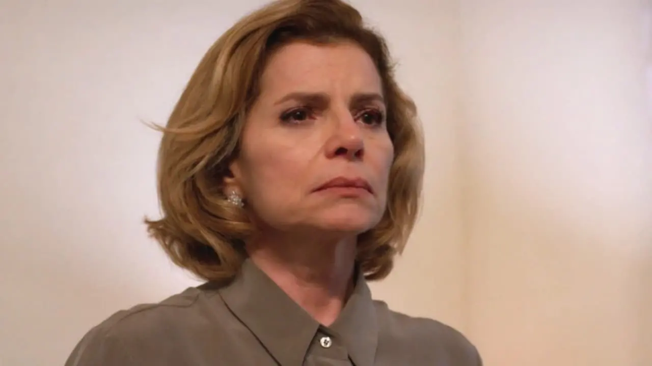 Mulher da terceira idade, branca, loira de cabelo curto e liso, com expressão triste. Usa terno cinza visível até o ombro, em cenário com fundo de parede branca.