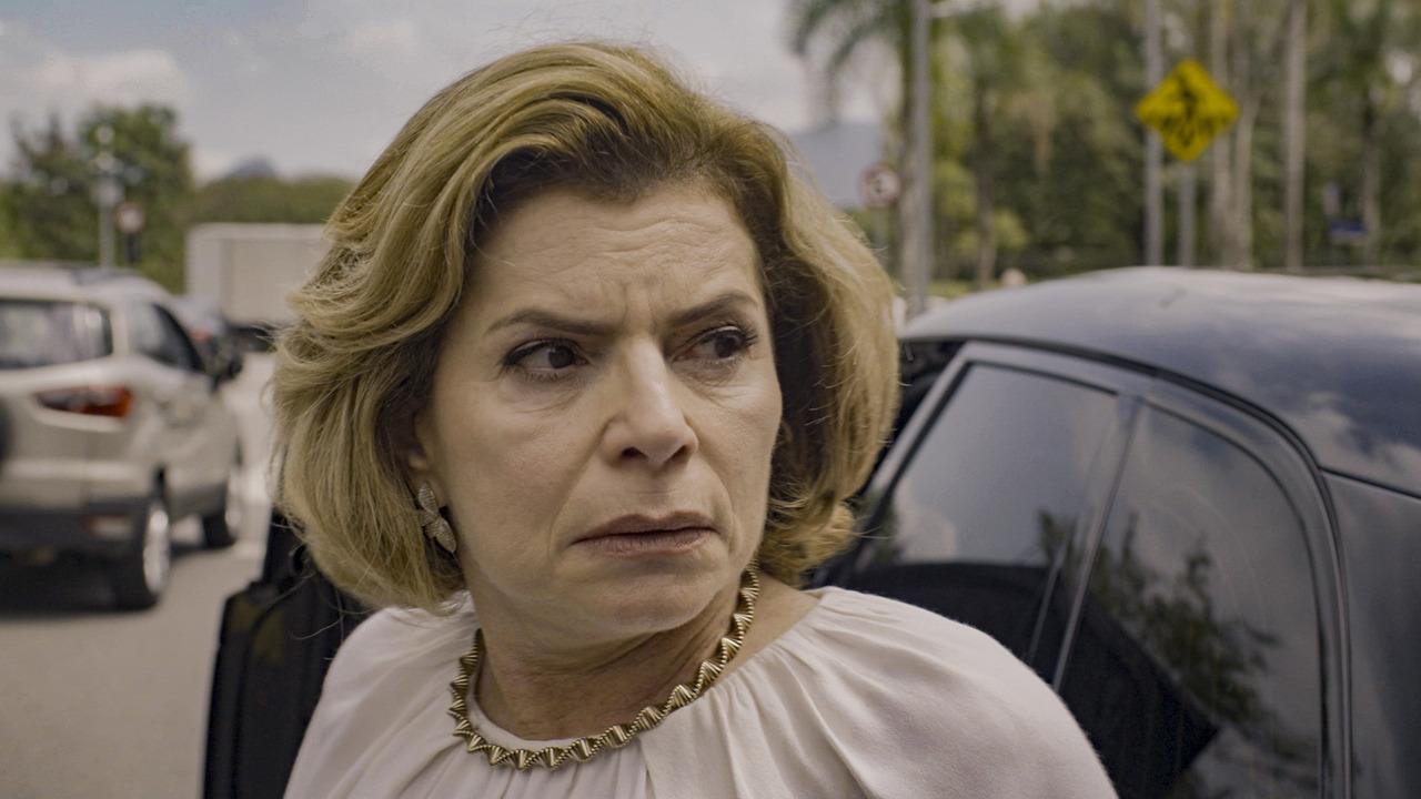 Mulher branca de cabelo loiro curto, da terceira idade, com blusa branca e colar dourado. Ao fundo, uma rua com um carro preto diretamente atrás dela e um carro cinza em segundo plano, na luz do dia, com algumas árvores atrás.