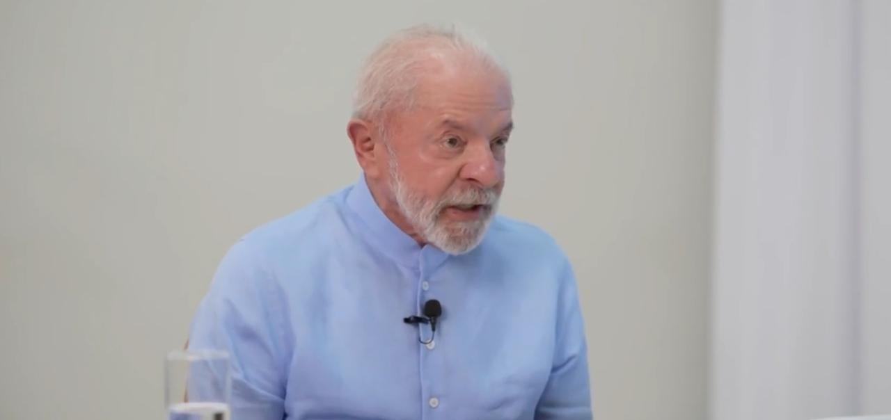 Lula, homem branco, idoso, de cabelo branco e ralo, barba e bigode brancos, em um fundo branco e vestindo uma blusa de manga longa azul clara.