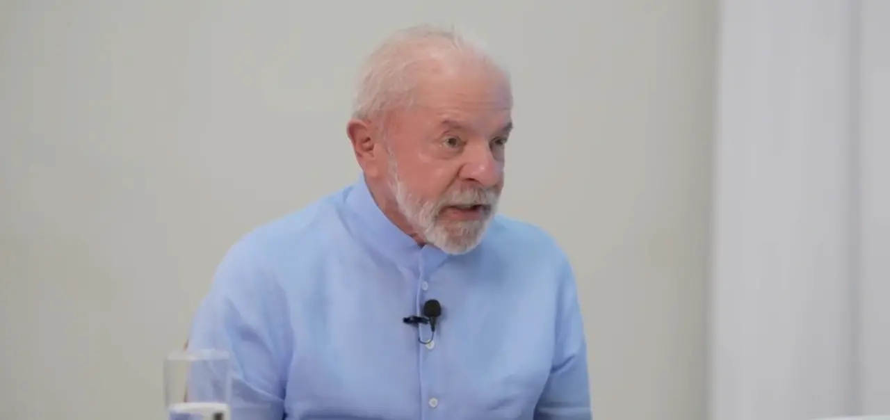 Lula, homem branco, idoso, de cabelo branco e ralo, barba e bigode brancos, em um fundo branco e vestindo uma blusa de manga longa azul clara.