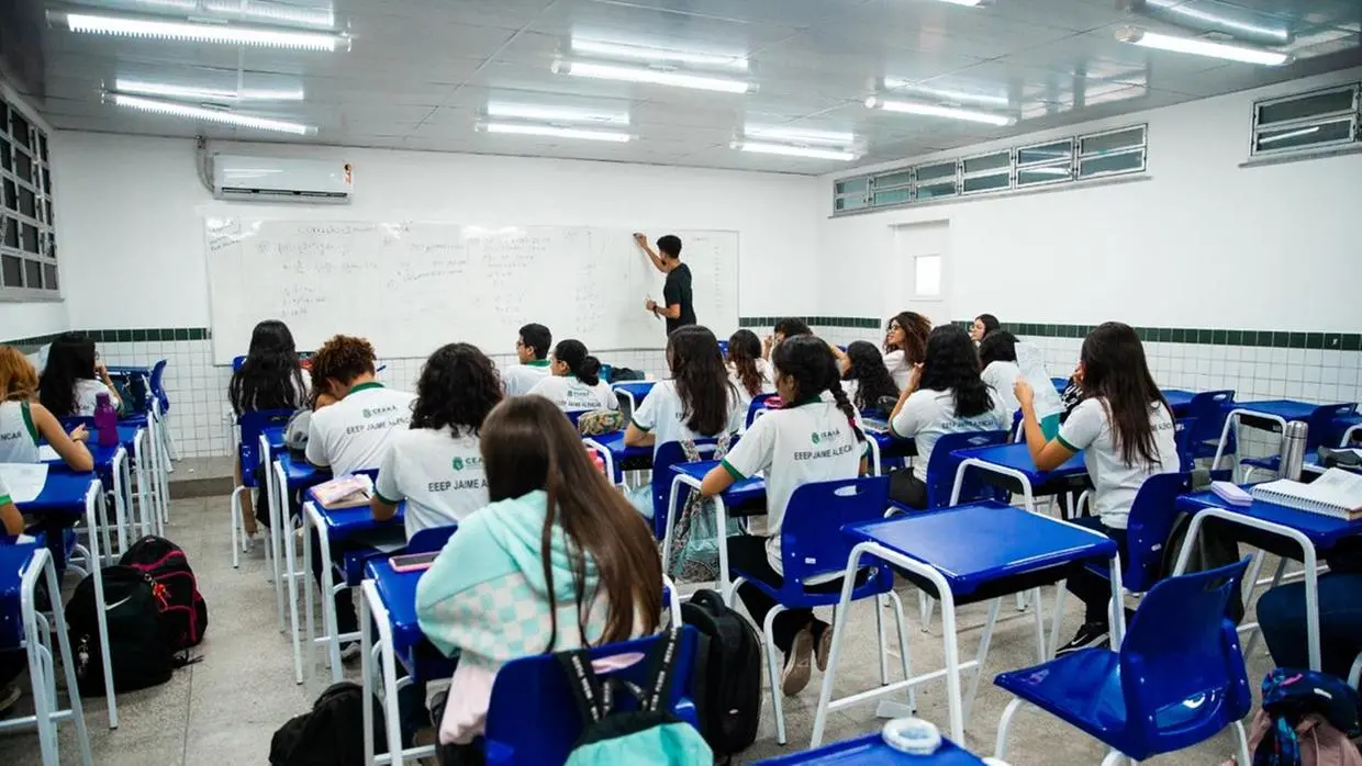 Sala de aula de escola pública no Ceará, com estudantes de uniforme branco e azul sentados em carteiras azuis, enquanto professor escreve no quadro branco.