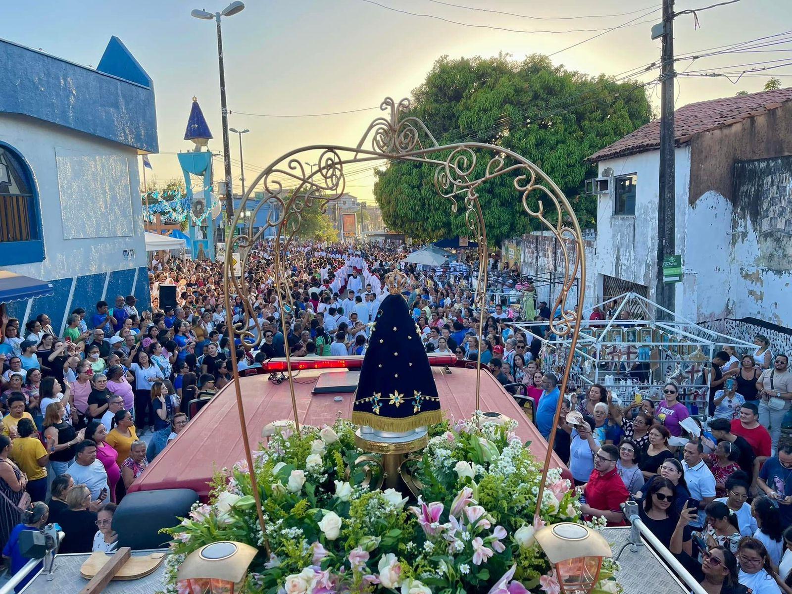 Grande multidão participa de celebração religiosa em uma rua movimentada, com destaque para uma procissão decorada com flores e uma imagem religiosa no centro.