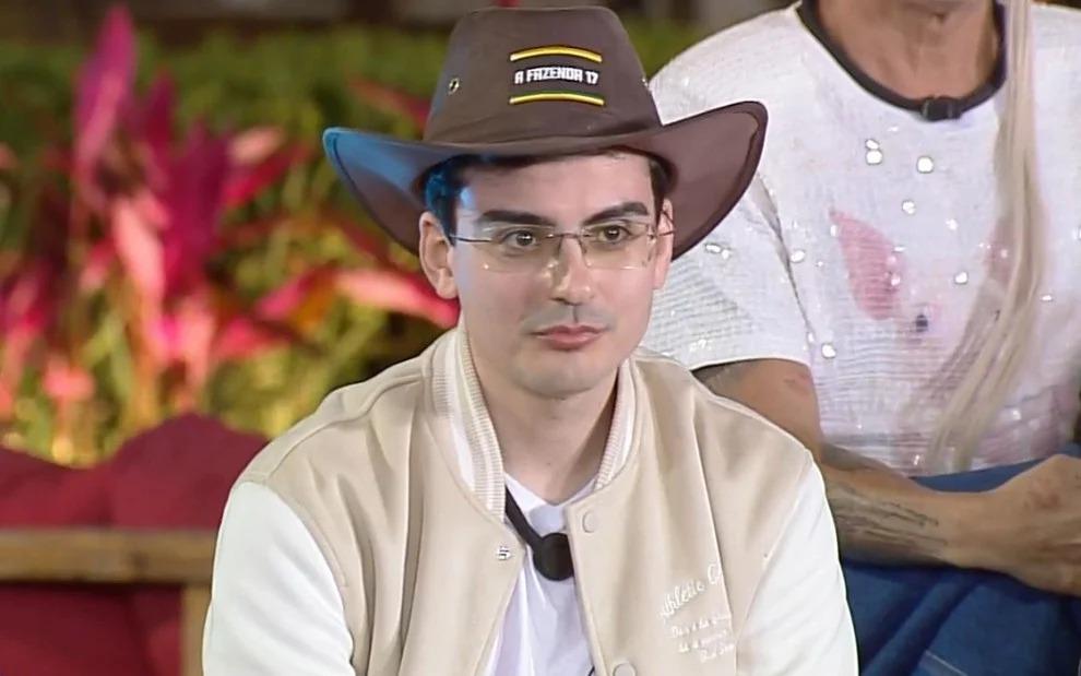 Dudu Camargo é um jovem branco de cabelos pretos e que usa óculos de grau. Na foto, ele está de chapéu de fazendeiro e usa uma jaqueta bege.
