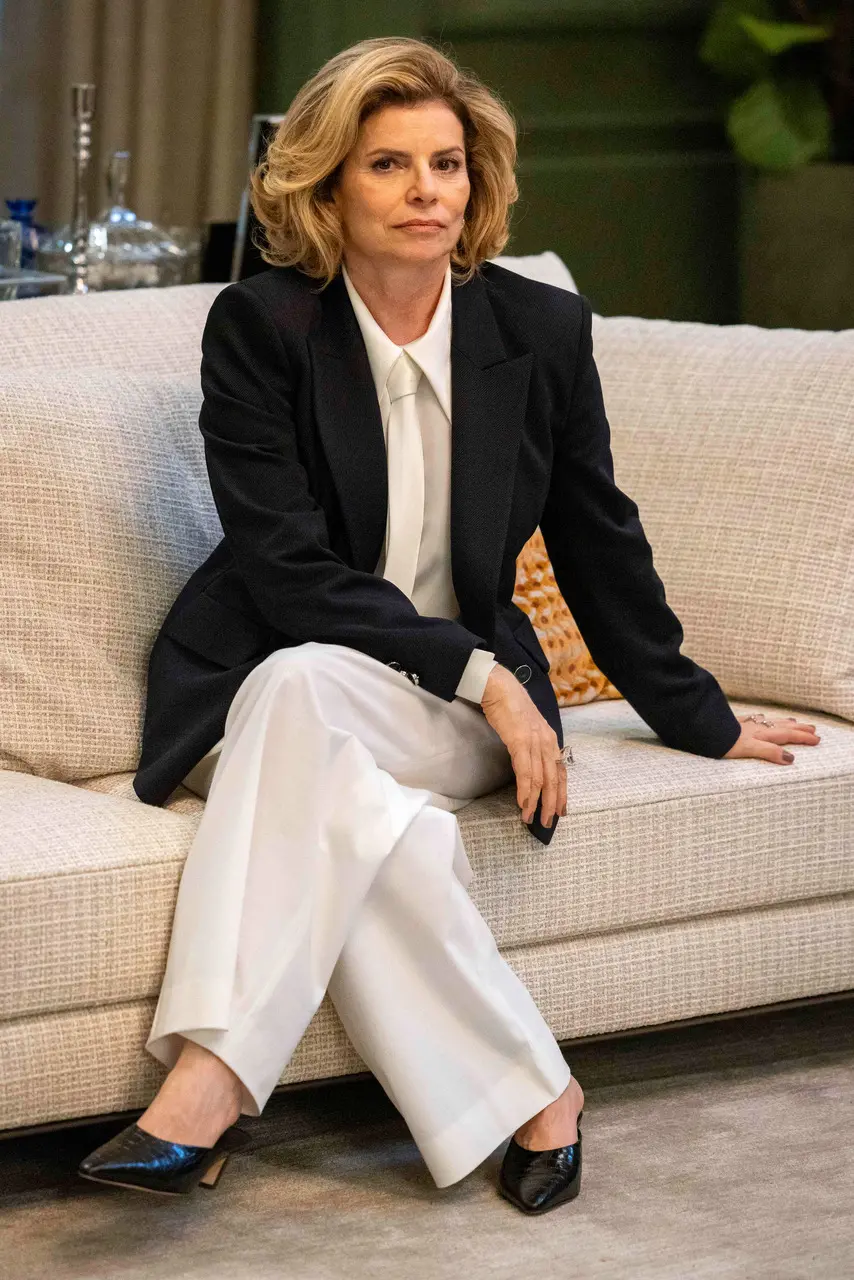 Personagem Odete Roitman, interpretada pela atriz Debora Bloch. Ela está sentada em um sofá branco, tem cabelos loiros lisos, na altura do ombro. Veste um blazer preto, com camisa, gravata e calça todos na cor branca.