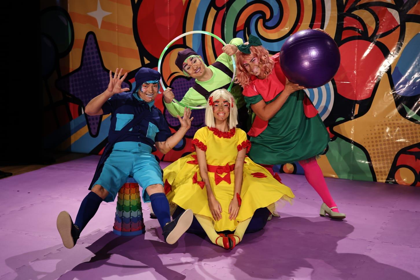 Na imagem, quatro atores em trajes coloridos e fantásticos, em uma performance teatral. Ao centro, sentada, está uma atriz de vestido amarelo e laço vermelho, com maquiagem branca e vermelha no rosto. À sua esquerda, um ator vestido de azul e turquesa, com maquiagem azul. Acima dela, uma atriz de verde-limão e um ator de camisa vermelha e saia verde-escura. Eles seguram aros e uma grande bola roxa. O fundo é vibrante com desenhos abstratos em cores brilhantes, incluindo estrelas. O piso é roxo. A imagem tem o crédito de Ares Soares.