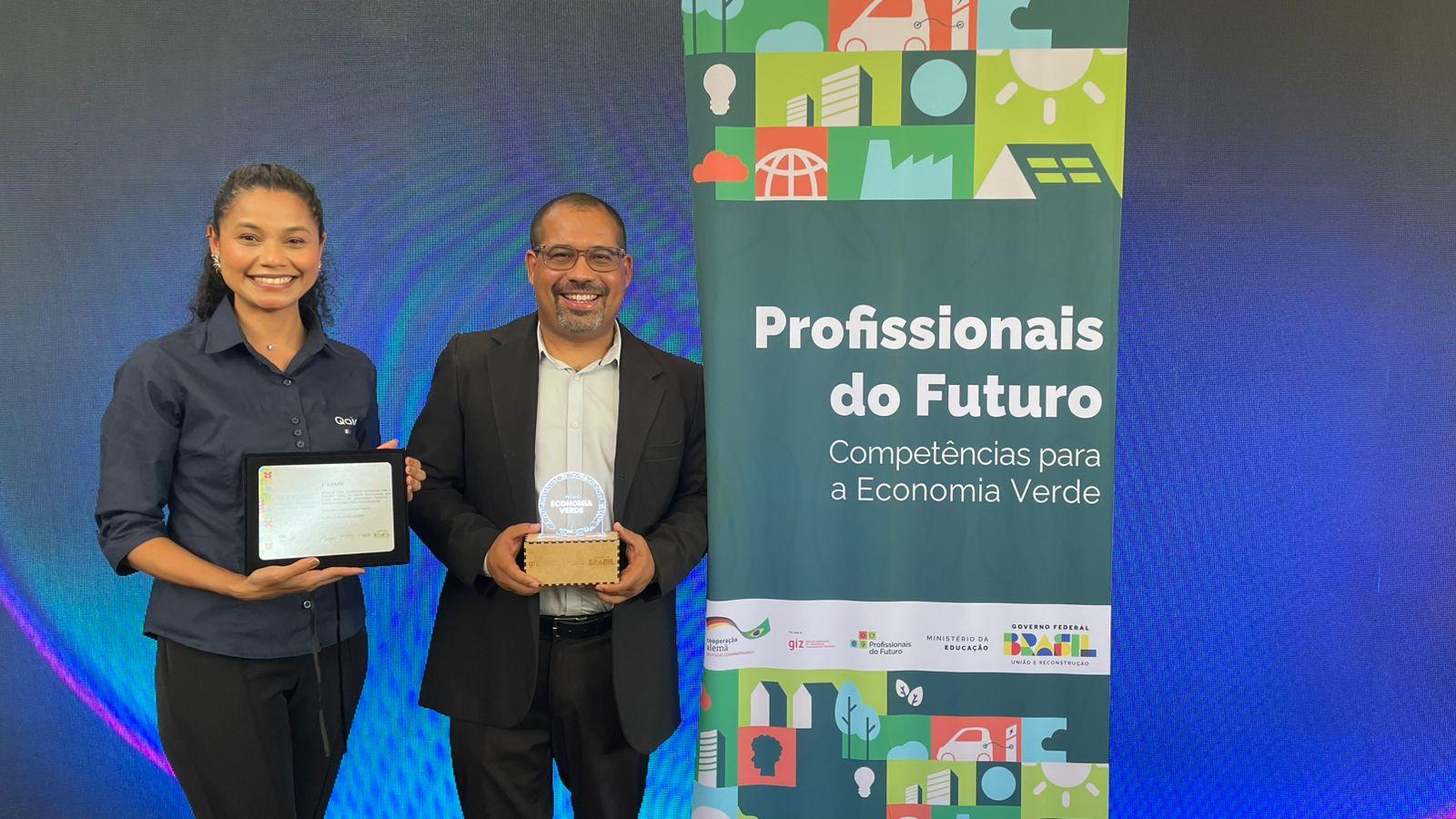 Mulher negra de cabelos cacheados segura placa e homem calvo de óculos e cavanhaque, de terno, segura troféu. Eles estão ao lado de placa do prêmio Profissionais do Futuro. Ao fundo, há um banner azul.