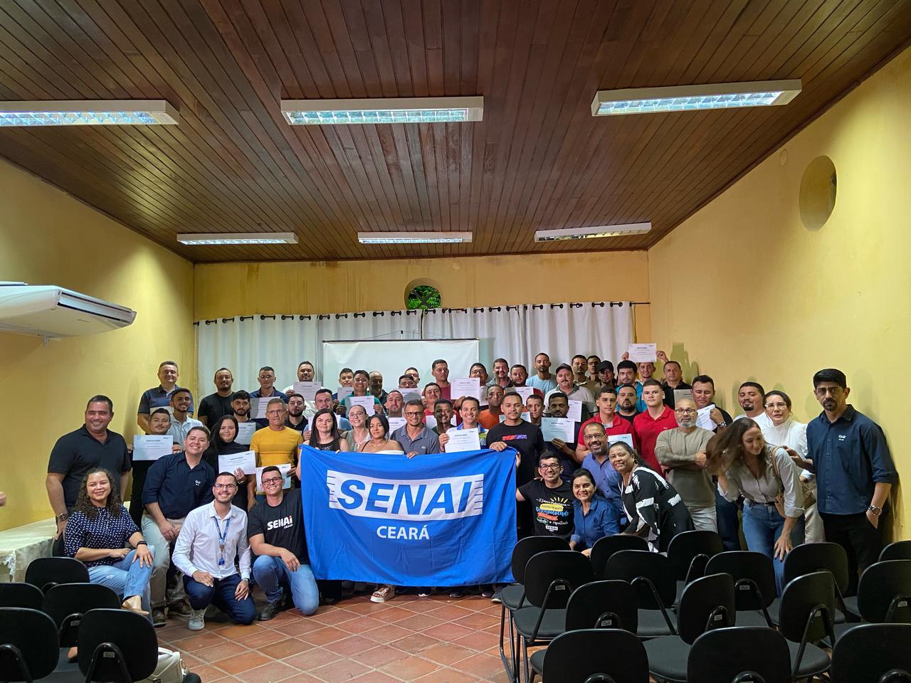 Foto de conclusão de turma de curso do Senai Ceará. Cerca de 50 pessoas se reúnem em auditório segurando certificados. Na frente, há uma bandeira com o nome da instituição.