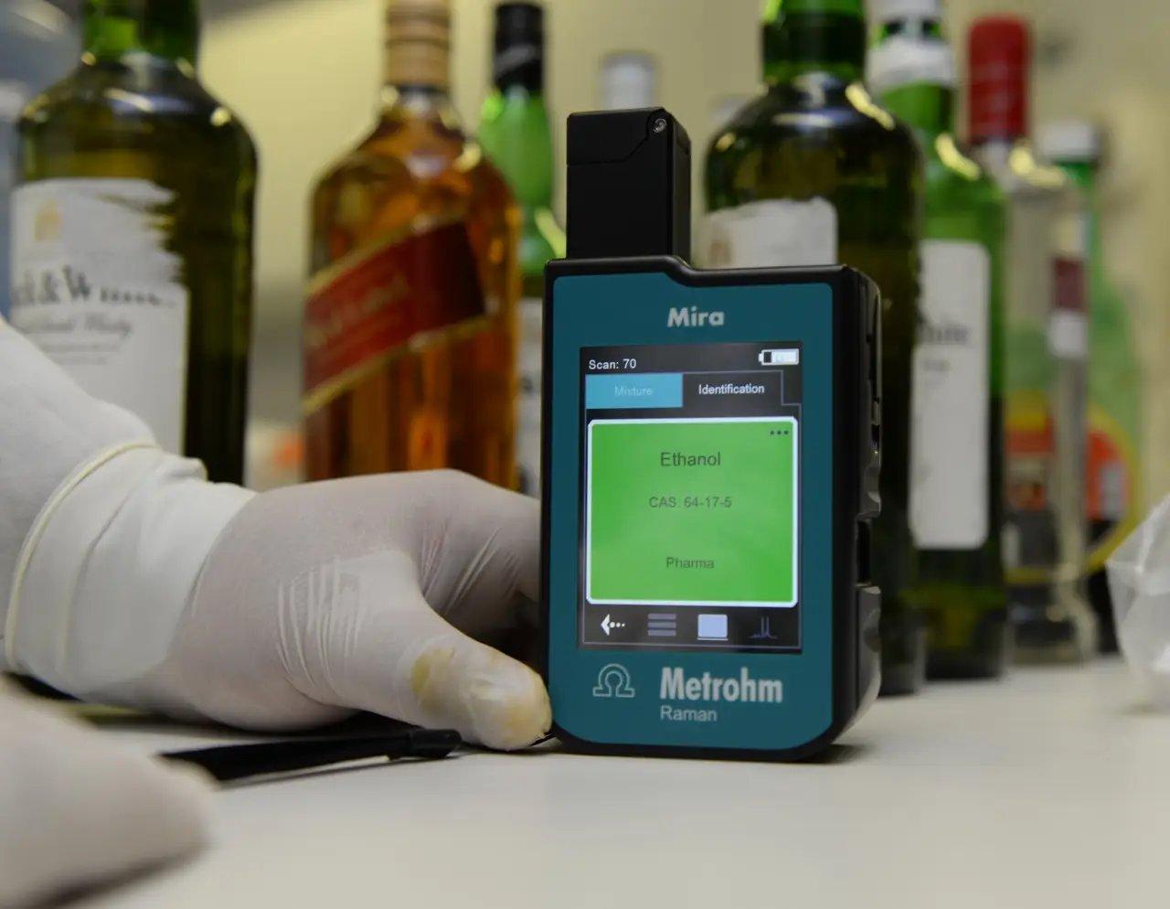 Mão com luva segura espectrômetro Raman portátil 'Metrohm Raman Mira', cuja tela exibe resultado de análise identificando 'Ethanol' (CAS 64-17-5) como substância presente. Ao fundo, há diversas garrafas de bebidas alcoólicas, indicando contexto de investigação ou verificação de conteúdo.