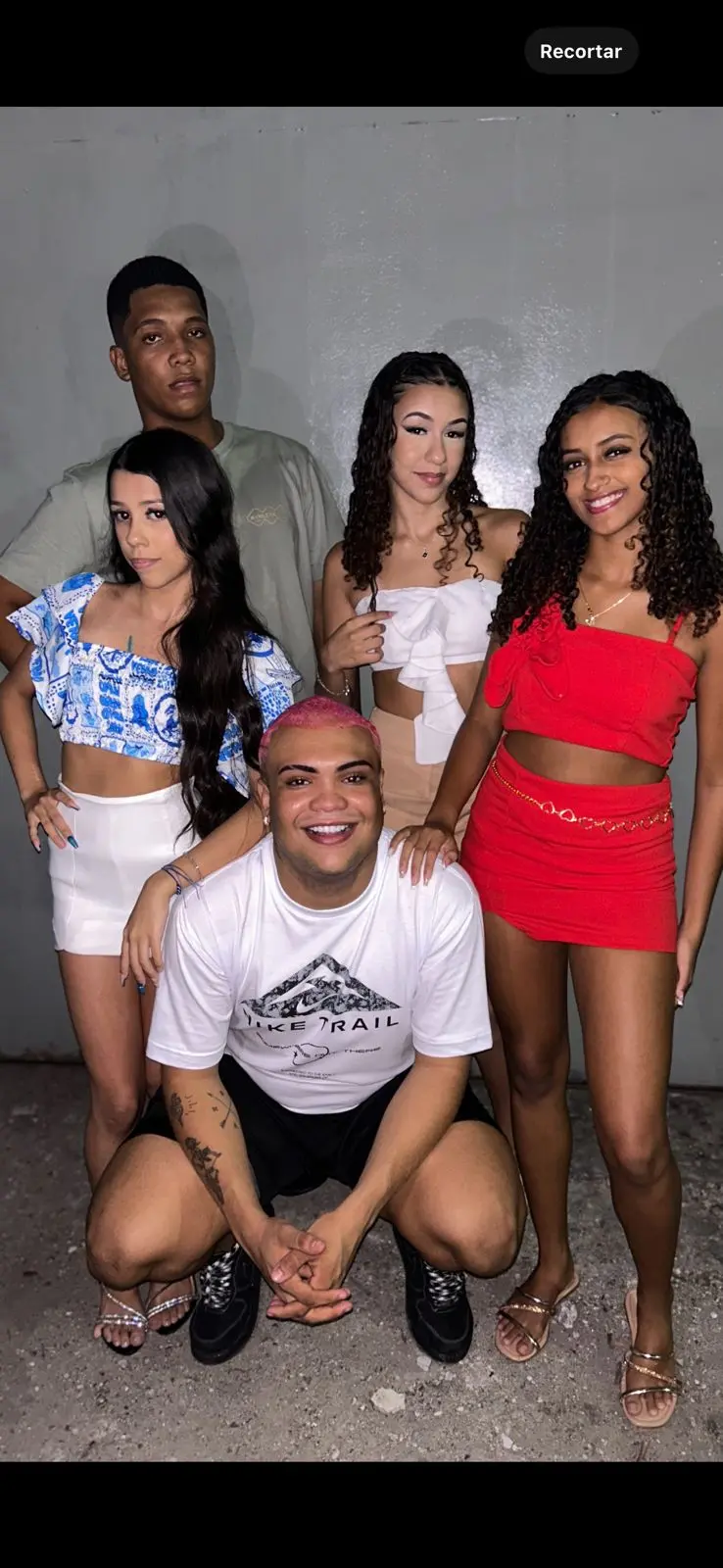 Cinco pessoas posam para foto em grupo: três em pé ao fundo e duas agachadas à frente. As roupas variam entre blusas e camisetas em tons de azul, branco, vermelho e cinza, combinadas com shorts e calças. A imagem parece capturar um momento descontraído entre amigos ou colegas.
