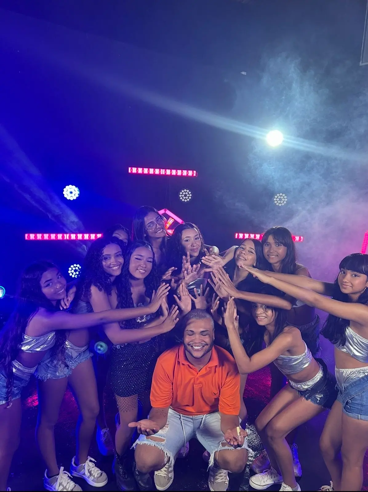 Grupo de doze pessoas posa em ambiente com iluminação colorida e efeitos de fumaça, sugerindo um estúdio ou palco. Onze estão em pé e uma pessoa ajoelhada ao centro, vestida com camisa laranja e jeans rasgados, recebe gestos de destaque dos demais. Os participantes usam roupas casuais e festivas, com tops brilhantes e shorts, em clima descontraído e alegre