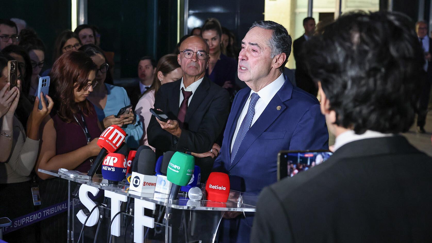 O ministro Luís Roberto Barroso falando em uma coletiva de imprensa, cercado por jornalistas e câmeras, em evento oficial
