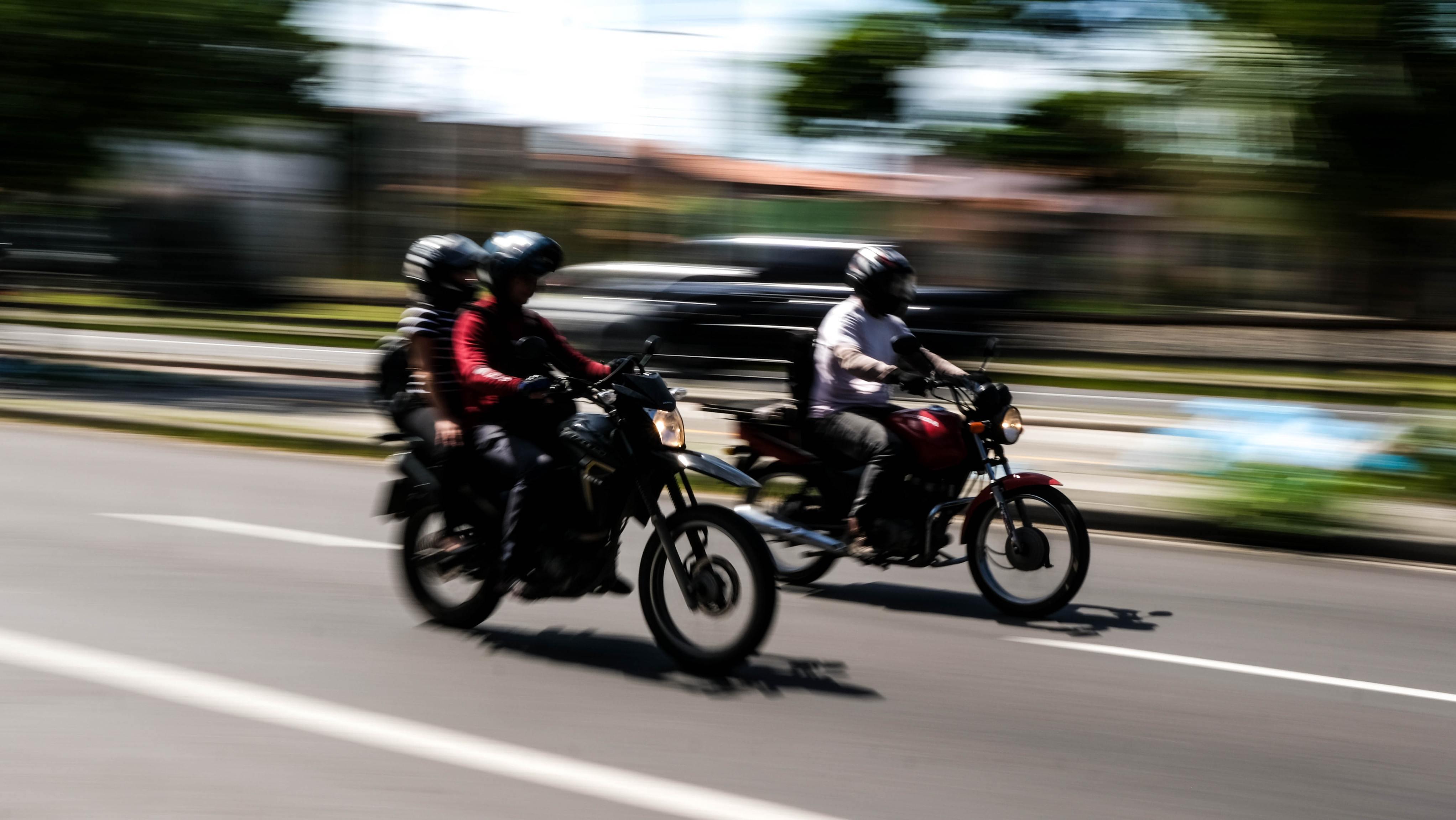 Duas motocicletas com condutores e passageiros de capacete em movimento rápido na rua. Fundo borrado para criar efeito de velocidade (panning).