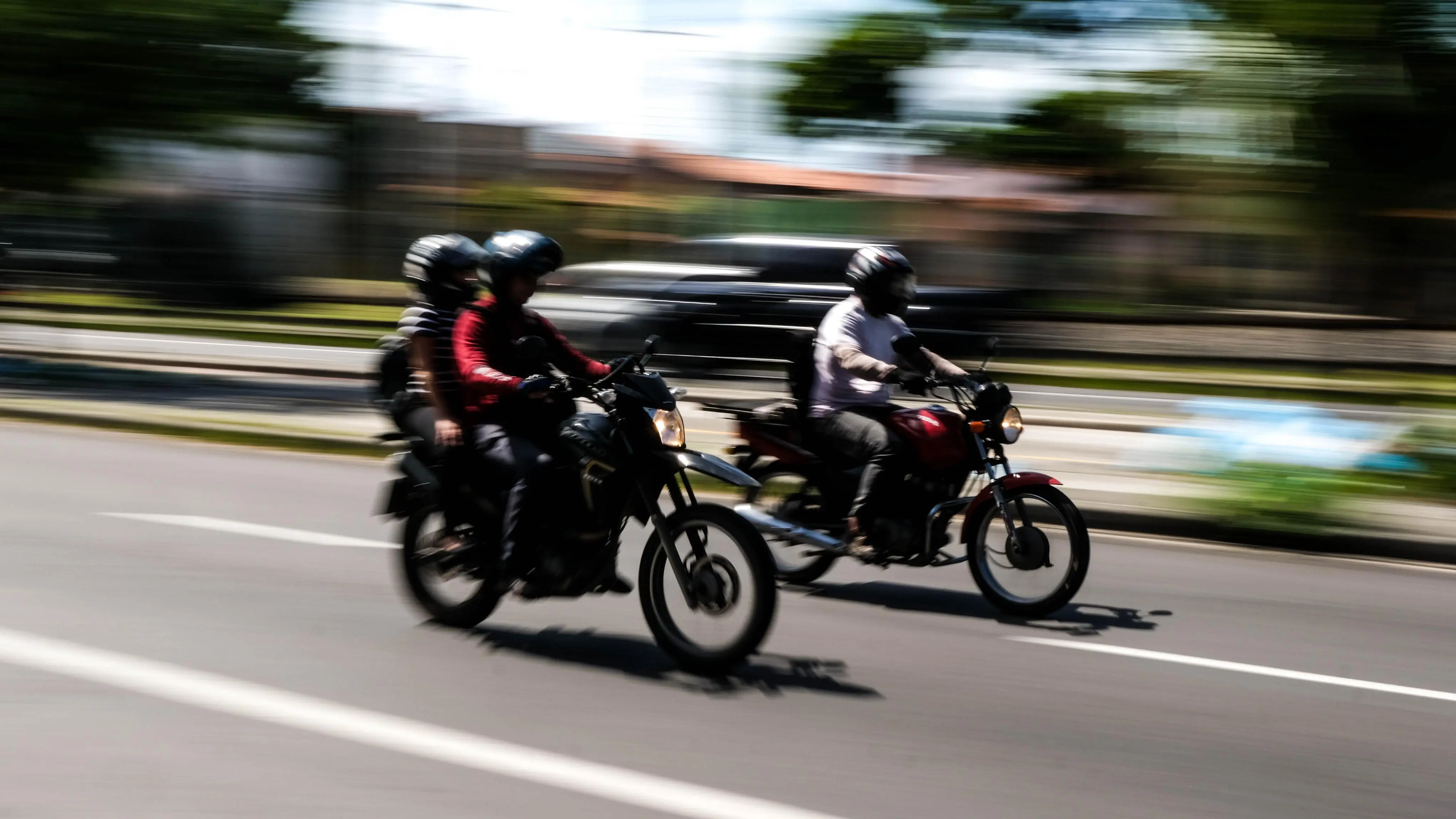 Duas motocicletas com condutores e passageiros de capacete em movimento rápido na rua. Fundo borrado para criar efeito de velocidade (panning).