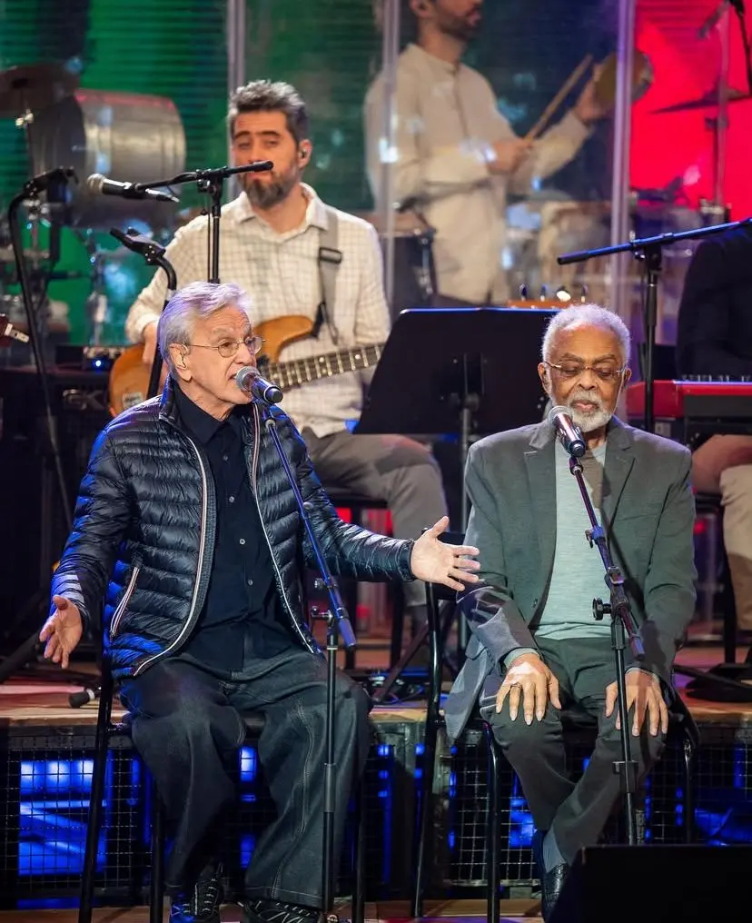 Show de música ao vivo com Caetano Veloso e Gilberto Gil cantando e tocando violão, acompanhado por músicos mais jovens ao fundo, em um palco iluminado. Em episódio especial em homenagem a Caetano Veloso e o aniversário de 25 anos do programa Altas Horas.