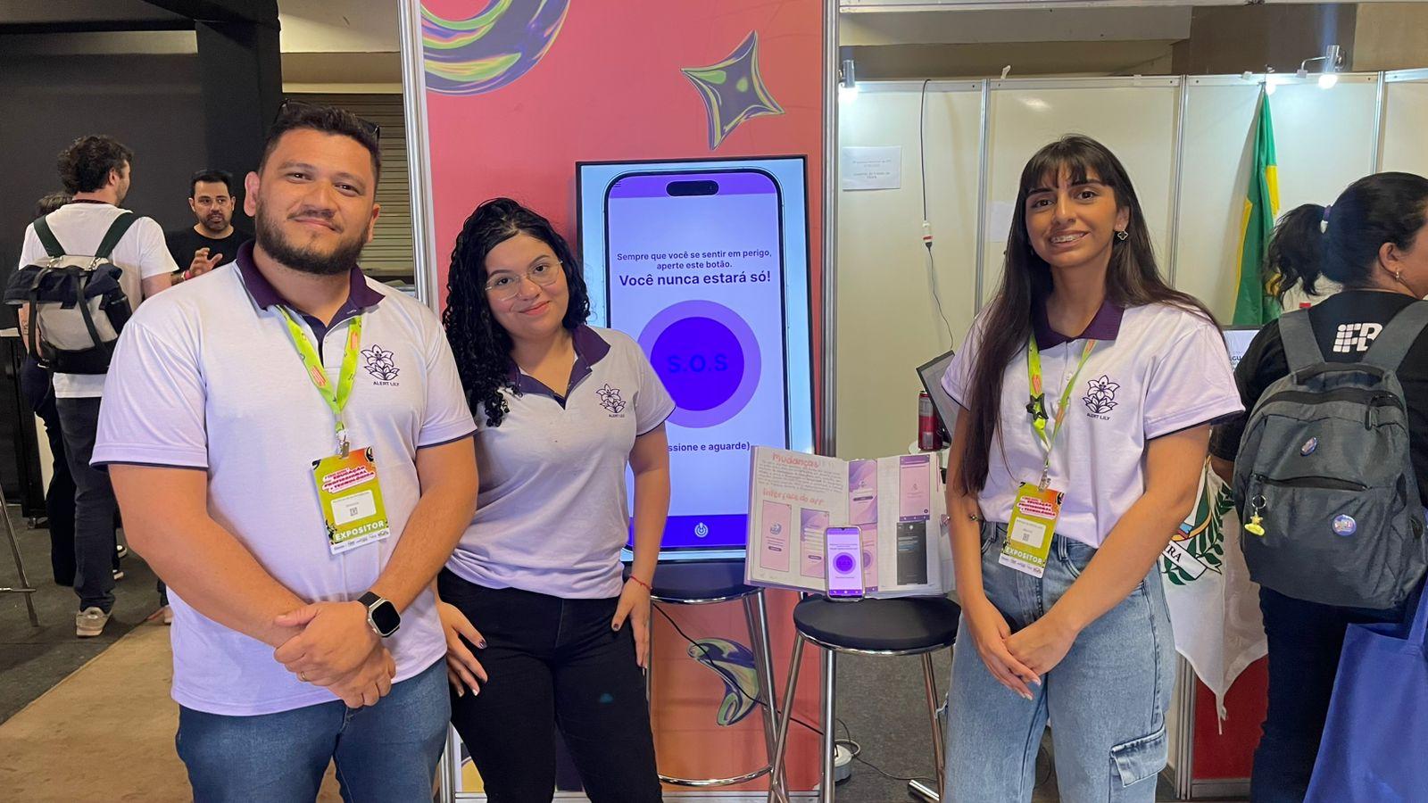 Professor e duas alunas com blusas de cor lilás posam para foto em frente a uma TV com a tela de um aplicativo de SOS para denunciar violência contra a mulher