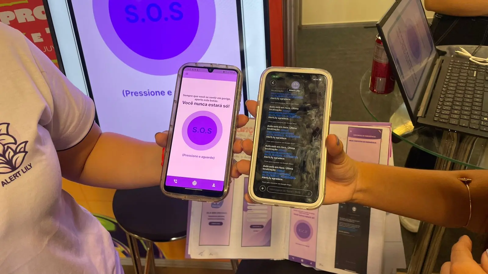 Foto detalhe de duas mãos segurando dois celulares. À esquerda, há um aplicativo com botão roxo escrito SOS. À direita, há uma página de mensagens com links para localizações por GPS.