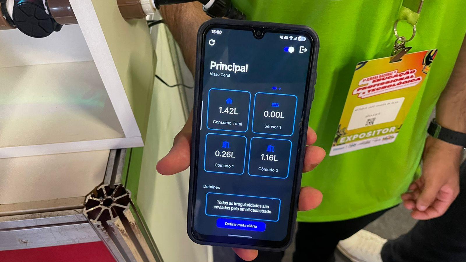 Mão segura e exibe a tela de um celular com um aplicativo de cor azul, mostrando diferentes dados sobre o consumo de água em uma casa. Pessoa que segura o aparelho usa blusa verde e um crachá