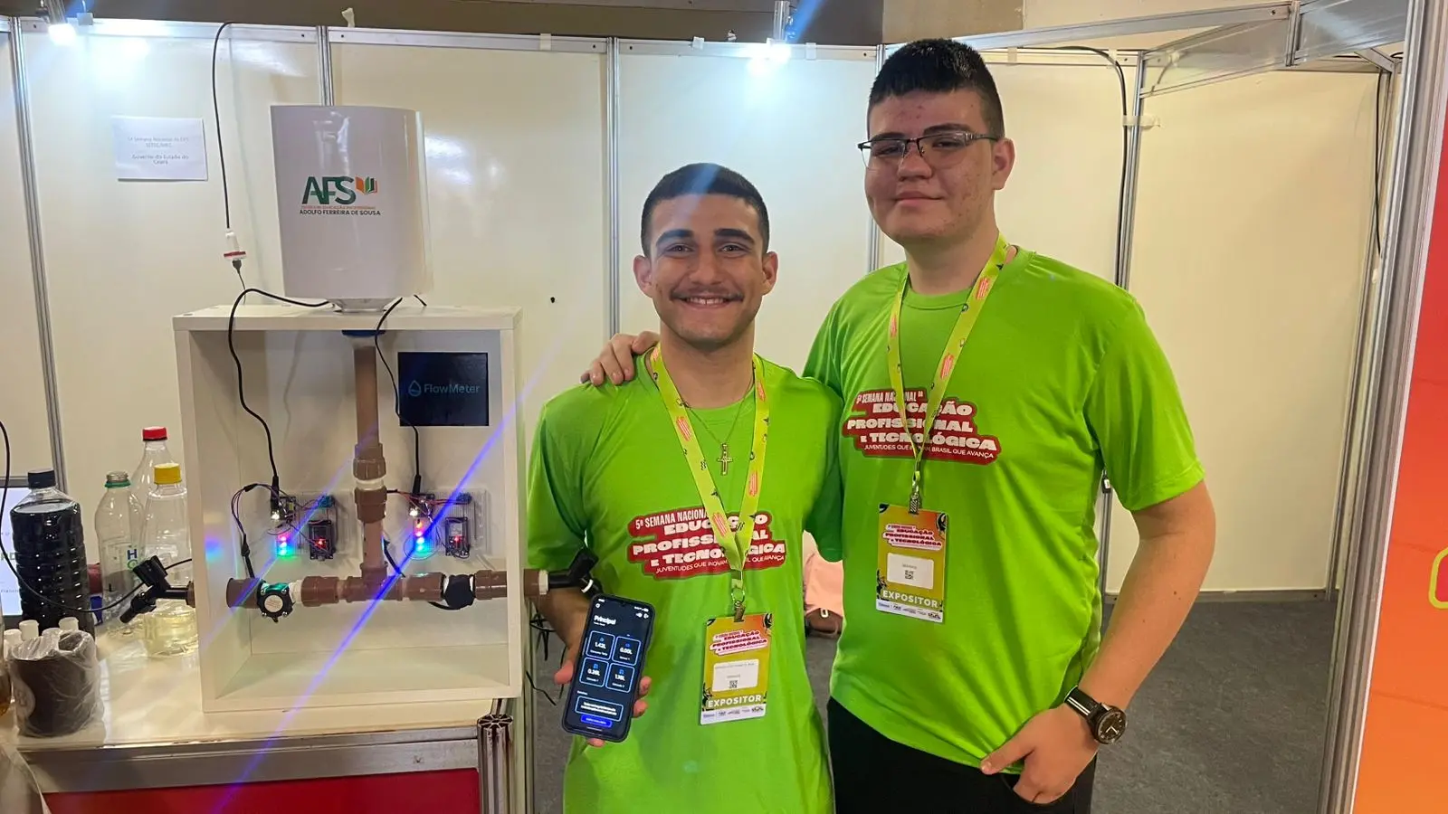 Dois alunos de blusa verde e crachá posam para foto segurando celular com aplicativo e maquete de monitoramento de água. Um é mais baixo e tem barba, o outro é mais alto e usa óculos