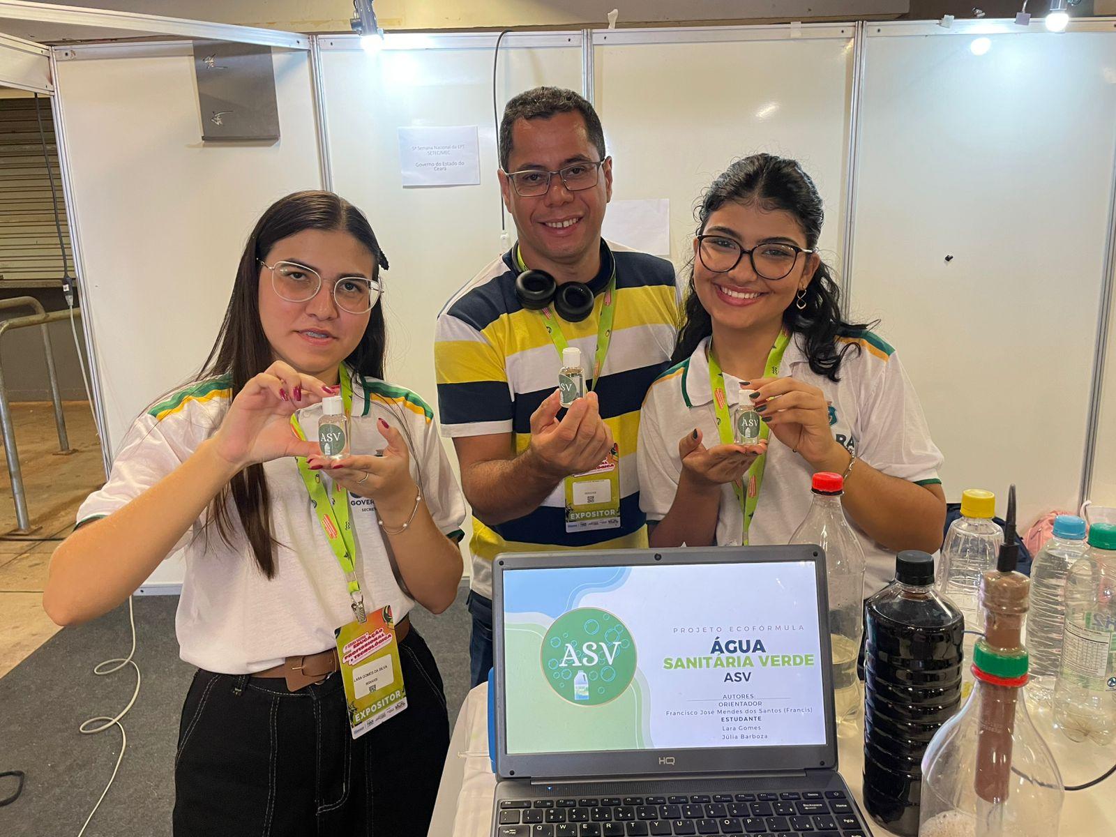 Duas alunas e um professor do Ceará posam para foto em estande de feira científica. Os três seguram frascos de água sanitária verde, projeto desenvolvido por eles. Numa mesinha à frente, há um notebook com detalhes do projeto e garrafas com substâncias usadas na produção.