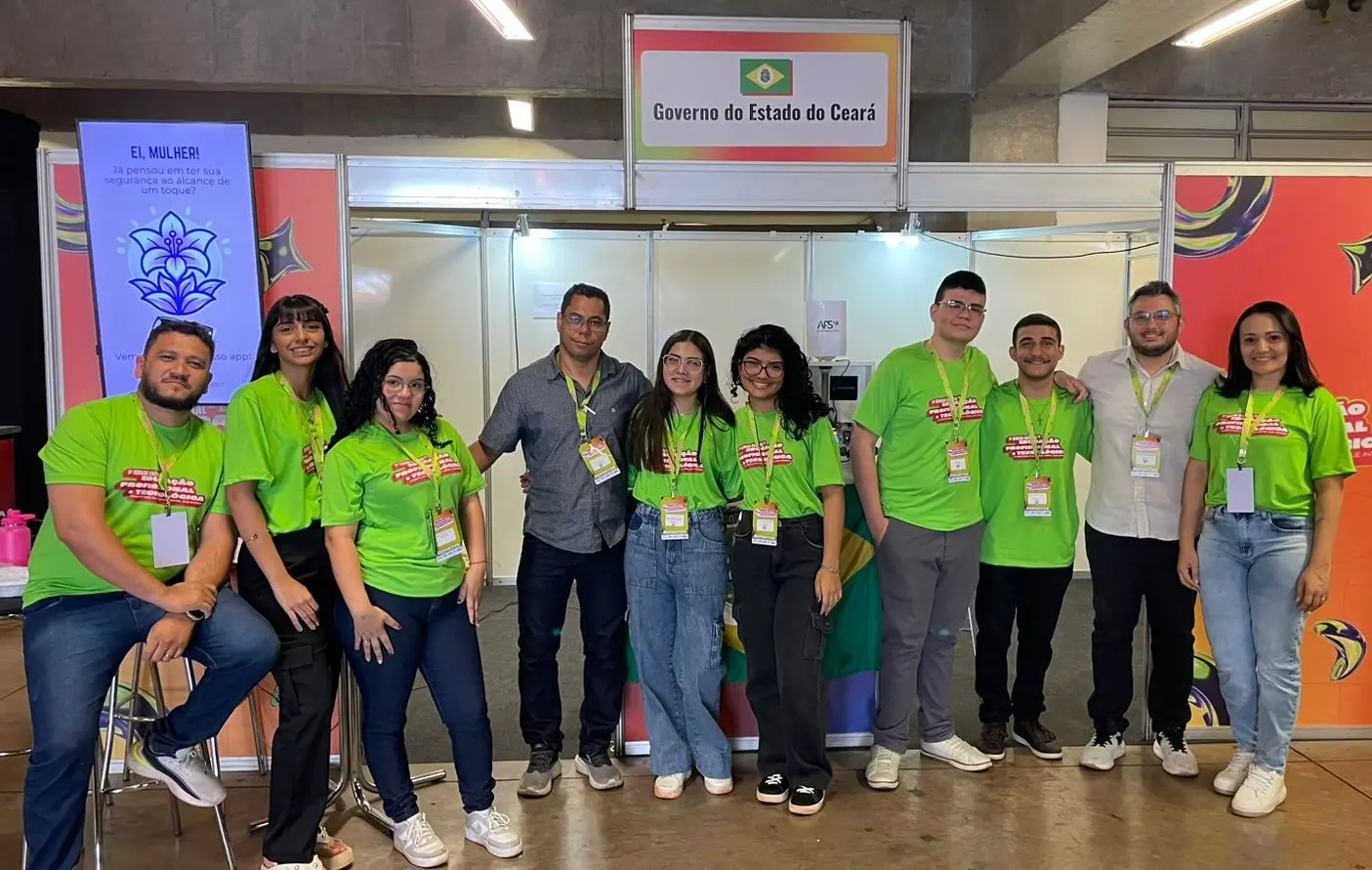 Foto com 10 pessoas com crachás, sendo seis alunos e quatro professores, posando para foto em frente a estande de feira científica representando o Estado do Ceará. Os alunos usam blusas verdes do evento.