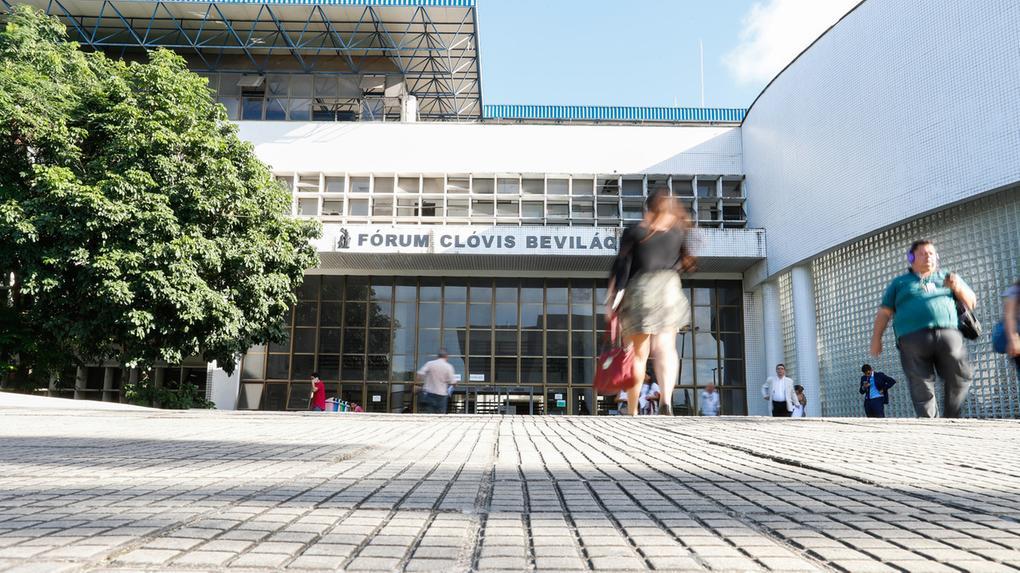 Foto mostra movimentação em frente ao Fórum Clóvis Bevilaqua, em Fortaleza. Em primeiro plano, é possível ver uma mulher de costas, blusa preta, saia e uma bolsa vermelha, e um homem de frente, com fone de ouvido, blusa azul e calça preta.