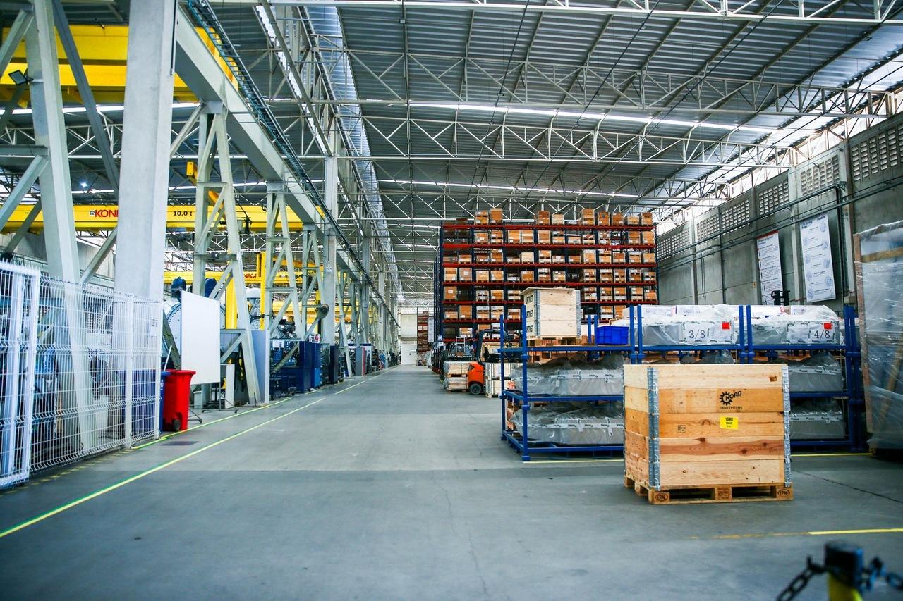 Imagem mostra interior de uma fábrica com estruturas metálicas e pilhas de produtos.