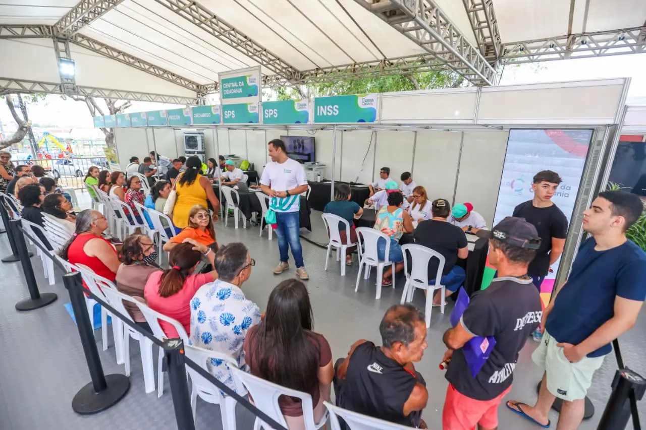 Pessoas aguardam atendimento em cadeiras sob uma tenda da Central da Cidadania, com guichês de órgãos públicos como SPS e INSS ao fundo, durante uma ação de serviços comunitários.