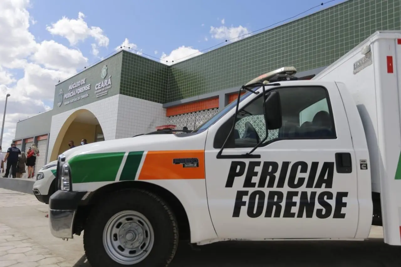 Na imagem, está uma viatura de cor branca da Perícia Forense do Ceará em frente a uma delegacia; é dia e está com bastante sol.