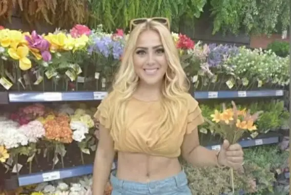 Foto que contém Ana Paula Veloso Fernandes, mulher acusada ser serial killer. Ela é loira, está com óculos escuros em cima da cabeça, veste uma camisa amarela escura e segura flores na mão esquerda.