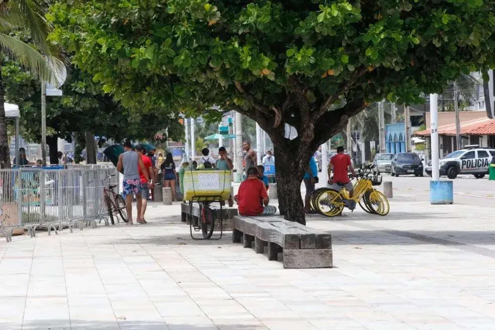 Foto aberta de calçadão na Avenida Beira-Mar com pedestres e vendedores com isopor. Ao centro, há uma árvore e um banco de cimento. Ao fundo, há um carro da Polícia Militar e lixeiras.