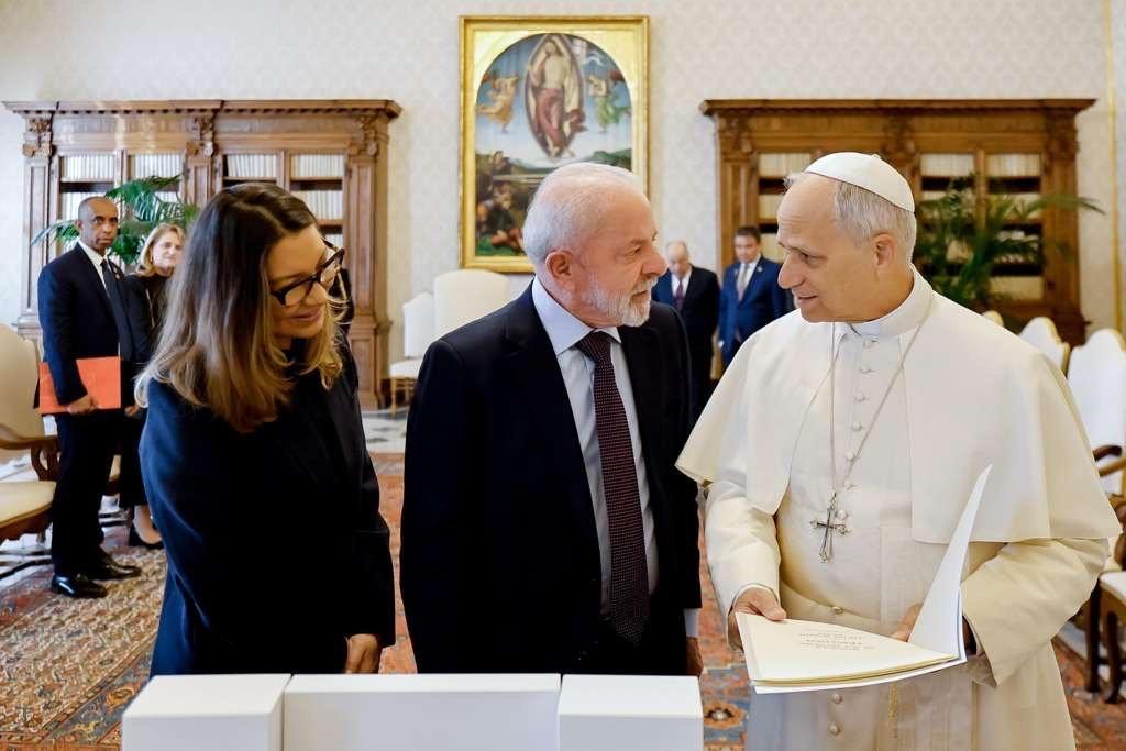 A imagem mostra o presidente Luiz Inácio Lula da Silva (Lula) e sua esposa, Janja, conversando com o Papa Francisco. Eles estão em uma audiência privada no Vaticano, com o Papa segurando um documento e falando com Lula, enquanto Janja observa. Ao fundo, é possível ver outras pessoas no ambiente, em uma sala com móveis clássicos e uma pintura religiosa na parede.