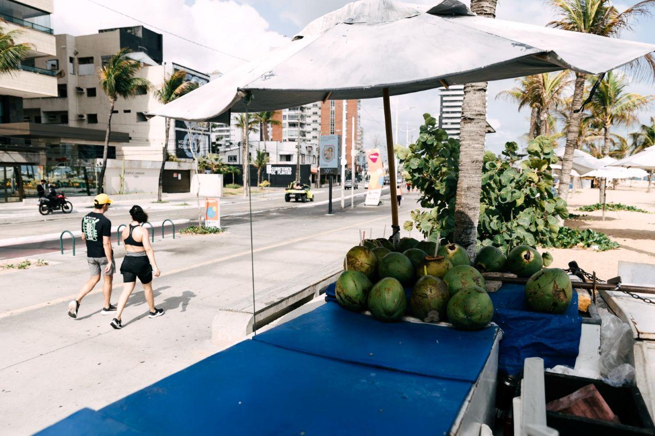 Imagem mostra calçadão da avenida beira-mar, em fortaleza. em destaque, imagem de quiosque que vende coco. ao fundo, na calçada, um homem e uma mulher estão caminhando.