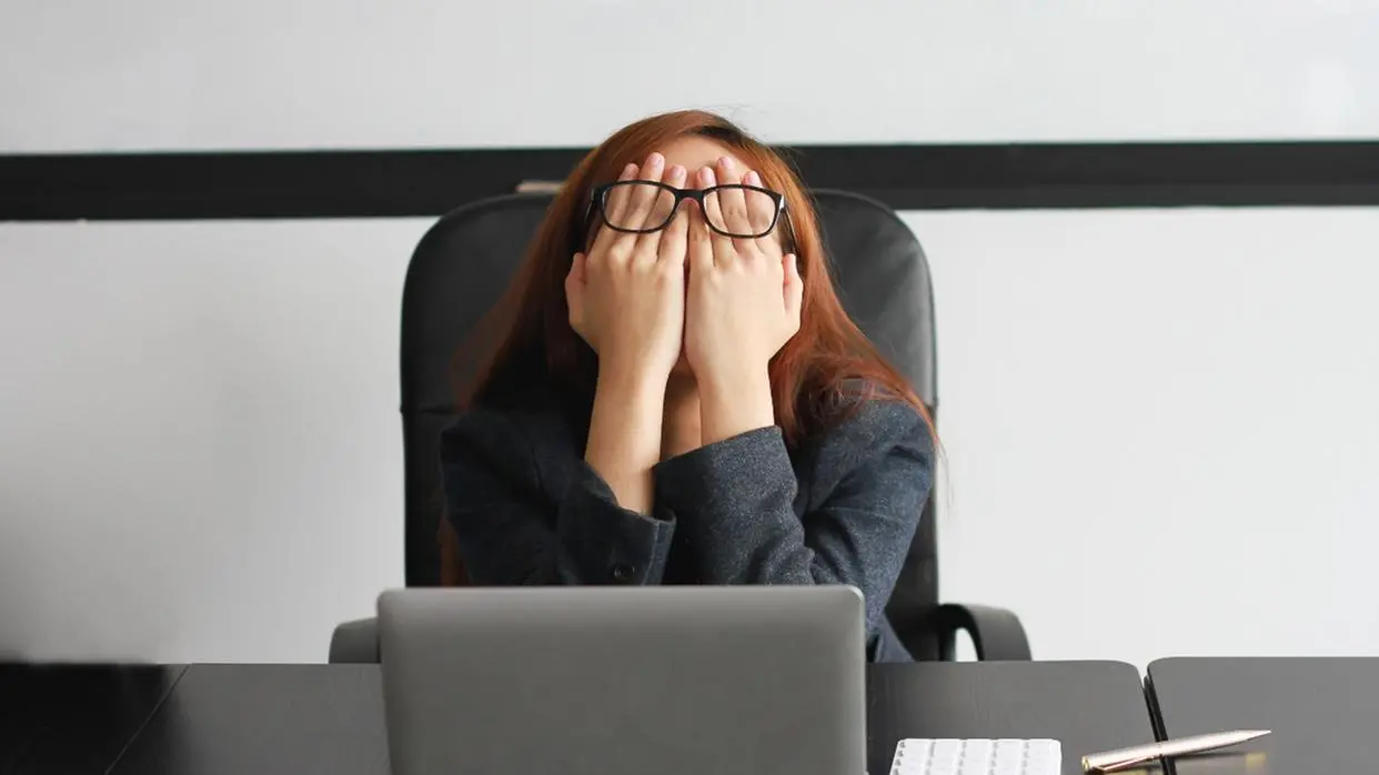 Imagem mostra uma mulher no ambiente de trabalho. Ela está com as mãos no rosto em sinal de estresse