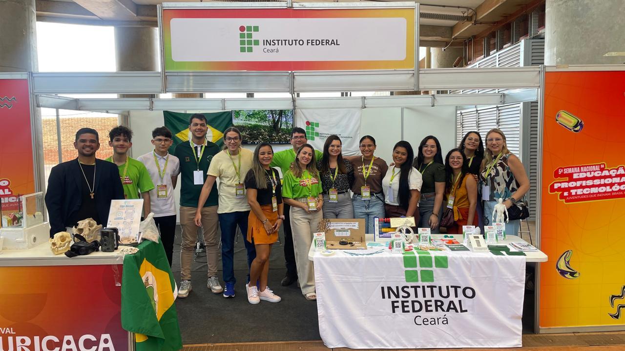 Grupo de alunos e professores do Ceará posam para foto em estande de feira de ciências em Brasília. Na frente deles, há uma bancada com a bandeira do Ceará. Na mesa ao lado, há objetos expostos sobre uma bandeira com a estampa do Instituto Federal do Educação do CE.