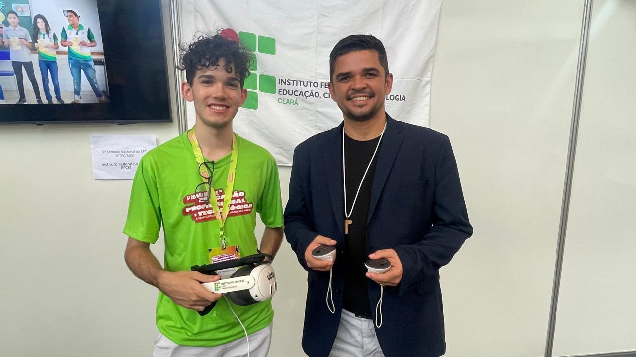 Um estudante e um professor mostram um dispositivo de realidade virtual desenvolvido no IFCE. O jovem segura o óculos com o logotipo do instituto, enquanto o professor exibe os controladores. O projeto foca em tecnologia imersiva aplicada à educação.