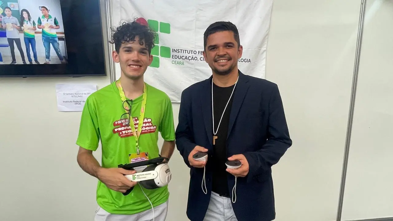 Um estudante e um professor mostram um dispositivo de realidade virtual desenvolvido no IFCE. O jovem segura o óculos com o logotipo do instituto, enquanto o professor exibe os controladores. O projeto foca em tecnologia imersiva aplicada à educação.