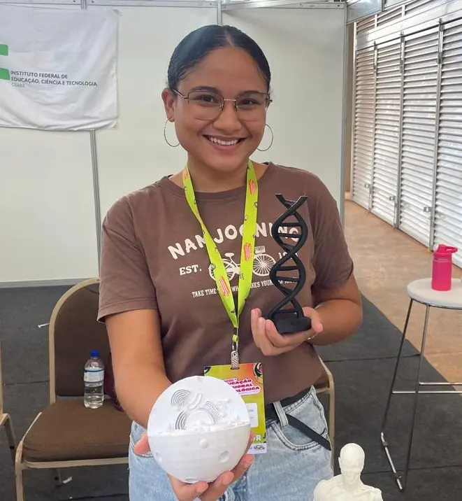 Uma estudante exibe modelos anatômicos e científicos impressos em 3D, incluindo uma hélice de DNA e uma esfera branca ilustrada. O projeto integra o ensino de biologia com tecnologia 3D. A aluna usa crachá de exposição e está em um estande do IFCE.