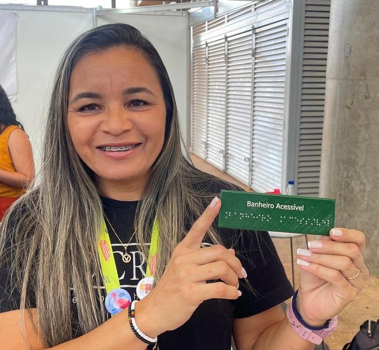 Uma mulher loira participante de feira científica segura uma placa verde com escrita em braille que indica “Banheiro Acessível”. Ela sorri e aponta para o item, destacando o foco em acessibilidade. No fundo, colegas interagem no estande do IFCE.